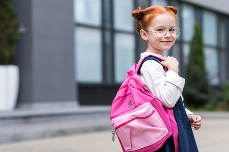 cute little redhead schoolgirl in eyeglasses holding backpack and smiling at camera の写真素材