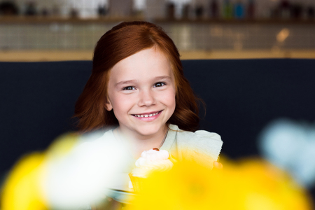 portrait of adorable happy redhead girl holding cupcake and smiling at camera in cafeの写真素材