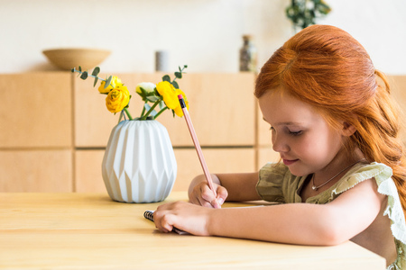 side view of cute redhead girl drawing with pencil while sitting at tableの写真素材