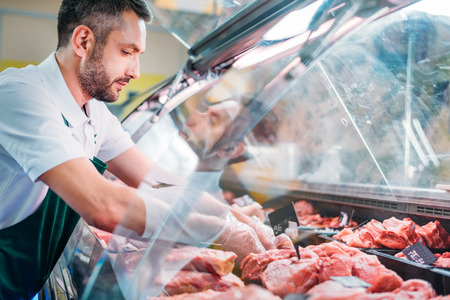 shop assistant in apron assorting fresh raw meat in supermarket