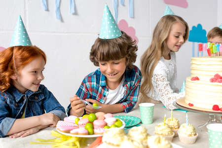 cute smiling kids eating sweets while sitting together at birthday tableの写真素材