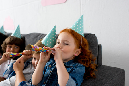 adorable little kids blowing in party blowers while sitting on sofa at birthday partyの写真素材