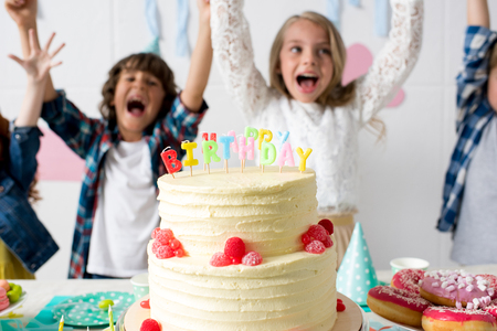 close-up view of delicious birthday cake on festive table and cheerful kids raising hands behindの写真素材