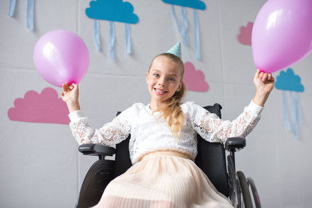 cheerful disabled girl holding balloons and smiling at camera at birthday partyの写真素材