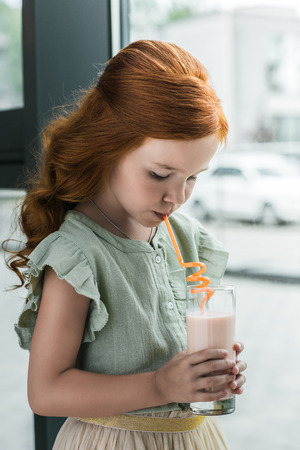 adorable little red haired girl drinking milkshake from glass in cafeの写真素材