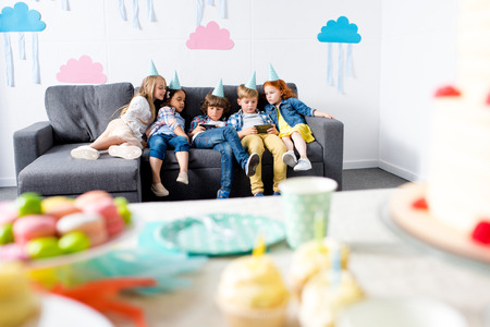 multiethnic kids using gadgets while sitting together on sofa at birthday partyの写真素材