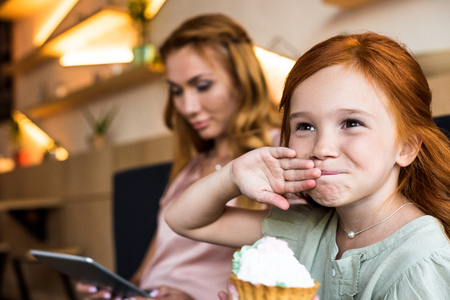 adorable small red haired girl eating cupcake while mother using digital tablet behindの写真素材