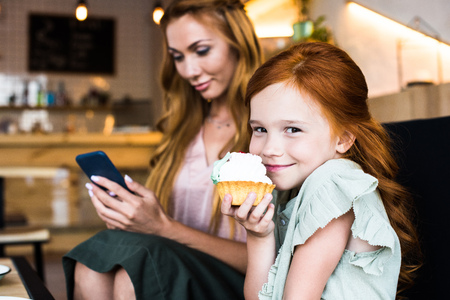 cute smiling redhead girl eating cupcake while mother using smartphone behindの写真素材