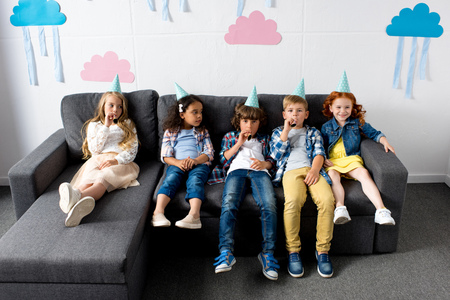 group of cute multiethnic kids in party caps sitting on sofa at birthday partyの写真素材