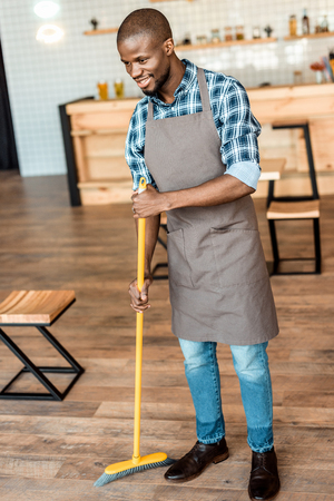 handsome smiling african american worker sweeping with broom in coffee shopの写真素材