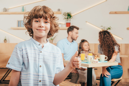 curly boy showing thumb up in cafe with his happy family blurred on backgroundの写真素材