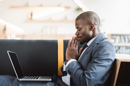 smiling african american businessman with closed eyes praying with laptopの写真素材