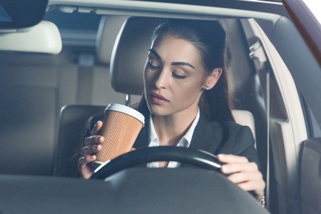 Young attractive woman in suit driving a car and drinking a cup of coffeeの写真素材