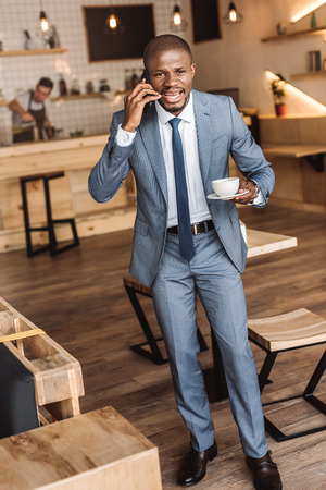handsome african american businessman holding cup of coffee and talking on smartphone in cafe の写真素材