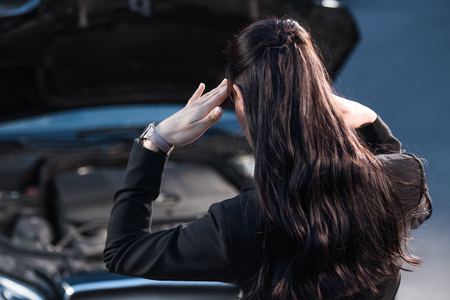 Young troubled woman rubbing temples while looking under hood of broken carの写真素材