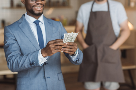 cropped view of handsome african american businessman holding and counting money, waiter standing behind in cafeの写真素材
