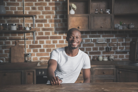 Portrait of young happy handsome african american man sitting at table in kitchenの写真素材