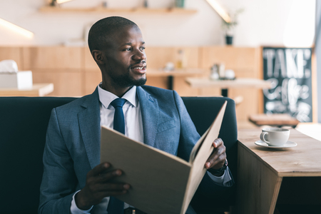 handsome african american businessman with cup of coffee reading menu in cafeの写真素材