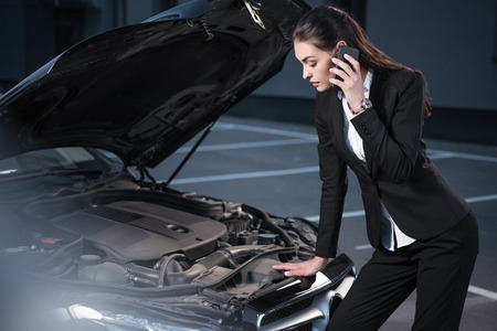 Young attractive woman in suit looking under hood of passenger car and talking on phoneの写真素材