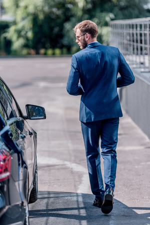 back view of stylish businessman in suit and sunglasses walking at carの写真素材
