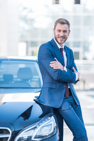 young smiling businessman in suit with crossed arms standing at car on parking  の写真素材