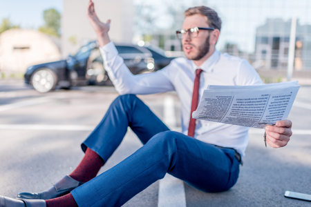 stressed businessman with newspaper sitting on parking with car behindの写真素材