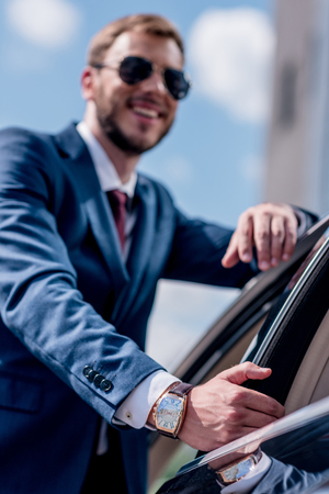 selective focus of young businessman in sunglasses with watch standing at car