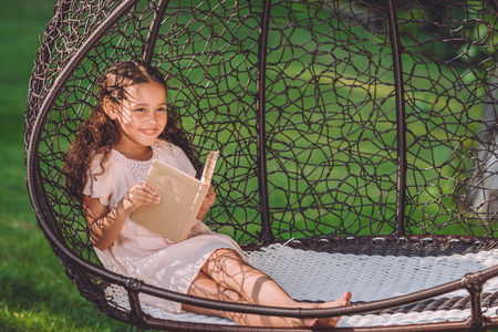 smiling african american girl reading book while sitting in swinging hanging chair in parkの写真素材