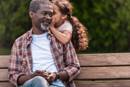 little adorable african american girl whispering to her grandfather while sitting on bench in park  の写真素材