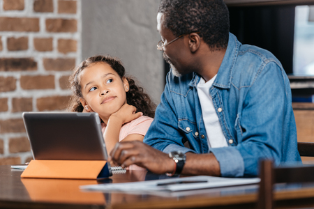 African american man sitting at table and using digital tablet with daughterの写真素材