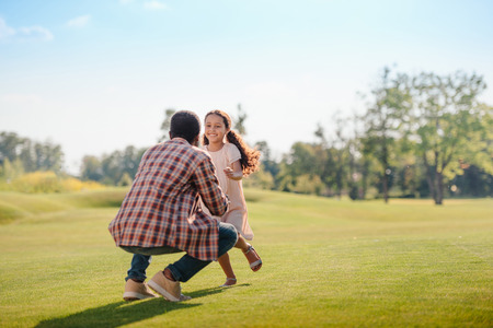 smiling african american granddaughter running to grandfather on green lawn in parkの写真素材