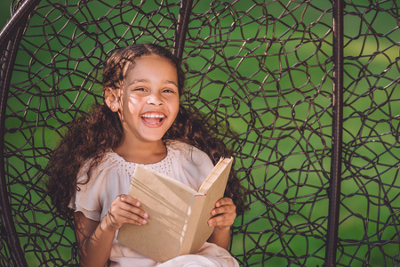 smiling african american girl reading book while sitting in swinging hanging chairの写真素材