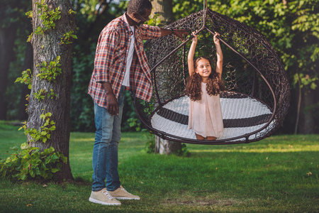 african american girl swinging on hanging chair while her grandfather standing near in parkの写真素材
