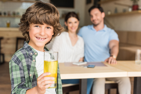 cute curly boy with glass of juice with his parents blurred on backgroundの写真素材