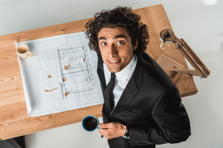 overhead view of smiling businessman with cup of coffee looking at camera while working with blueprints at workplaceの写真素材