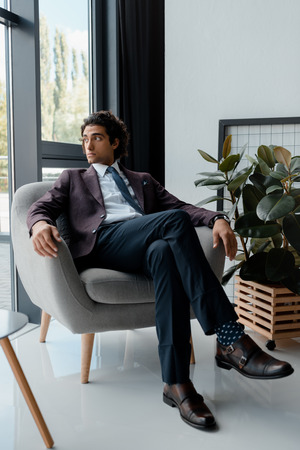 businessman in suit looking out window while resting in armchair in officeの写真素材
