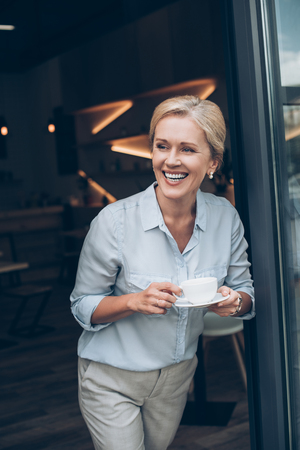 portrait of happy mature woman with cup of coffee in hands looking out coffee shopの写真素材