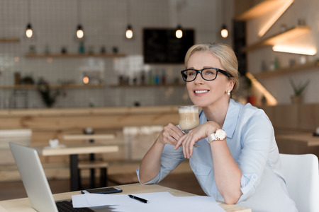 smiling mature businesswoman in eyeglasses drinking coffee while working in cafeの写真素材