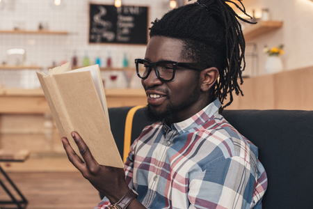 smiling african american man reading book in coffee shopの写真素材