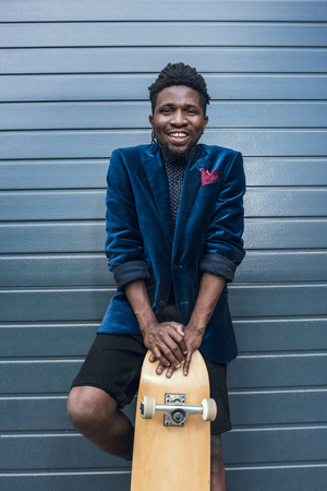 smiling stylish african american man in blue jacket leaning on skateboardの写真素材