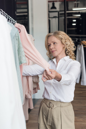 portrait of concentrated mature woman choosing clothes in clothing storeの写真素材