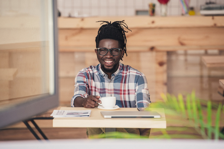 cheerful african american man drinking coffee at table in cafeの写真素材