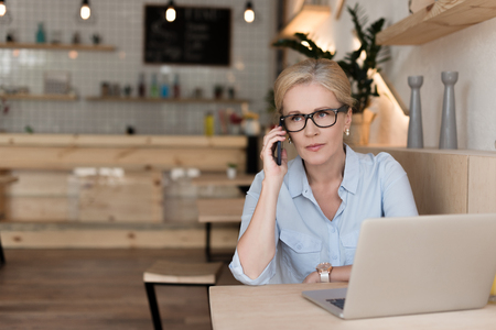 serious mature businesswoman in eyeglasses talking on smartphone and using laptop in cafeの写真素材