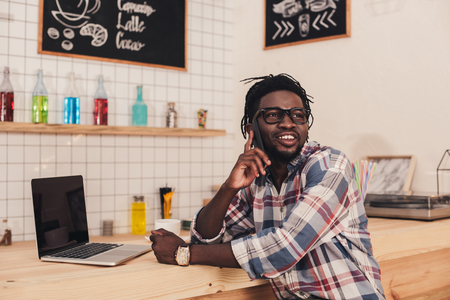 smiling african american man talking on smartphone and using laptop at bar counter in cafeの写真素材