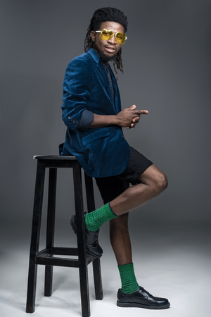 stylish african american man in blue jacket leaning on chair on gray の写真素材
