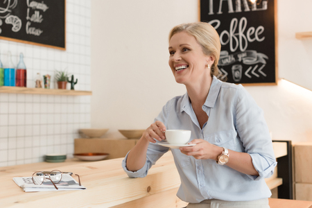 cheerful mature woman laughing while drinking coffee and looking away in cafeの写真素材