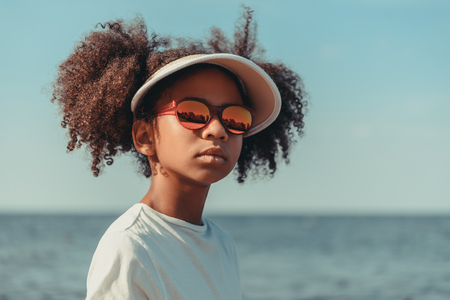 beautiful african american girl in cap and sunglasses looking away while standing on beachの写真素材