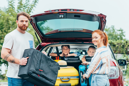 young parents packing luggage in trunk of car with kids looking from backseatsの写真素材
