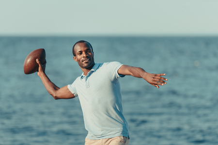 handsome young african american man throwing rugby ball on beachの写真素材