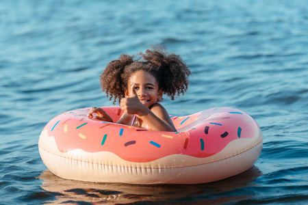 cute smiling african american girl swimming in rubber ring and showing thumb upの写真素材